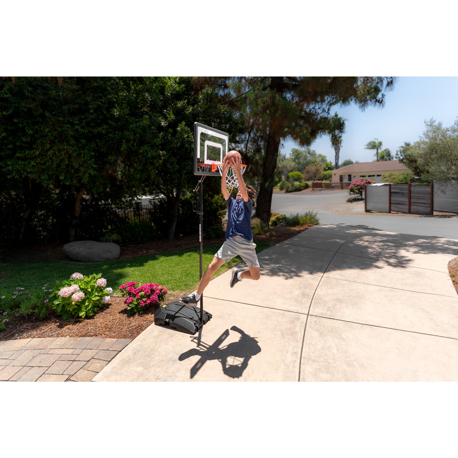 Boy jumps and shoots basketball outdoors near a portable hoop in a residential yard