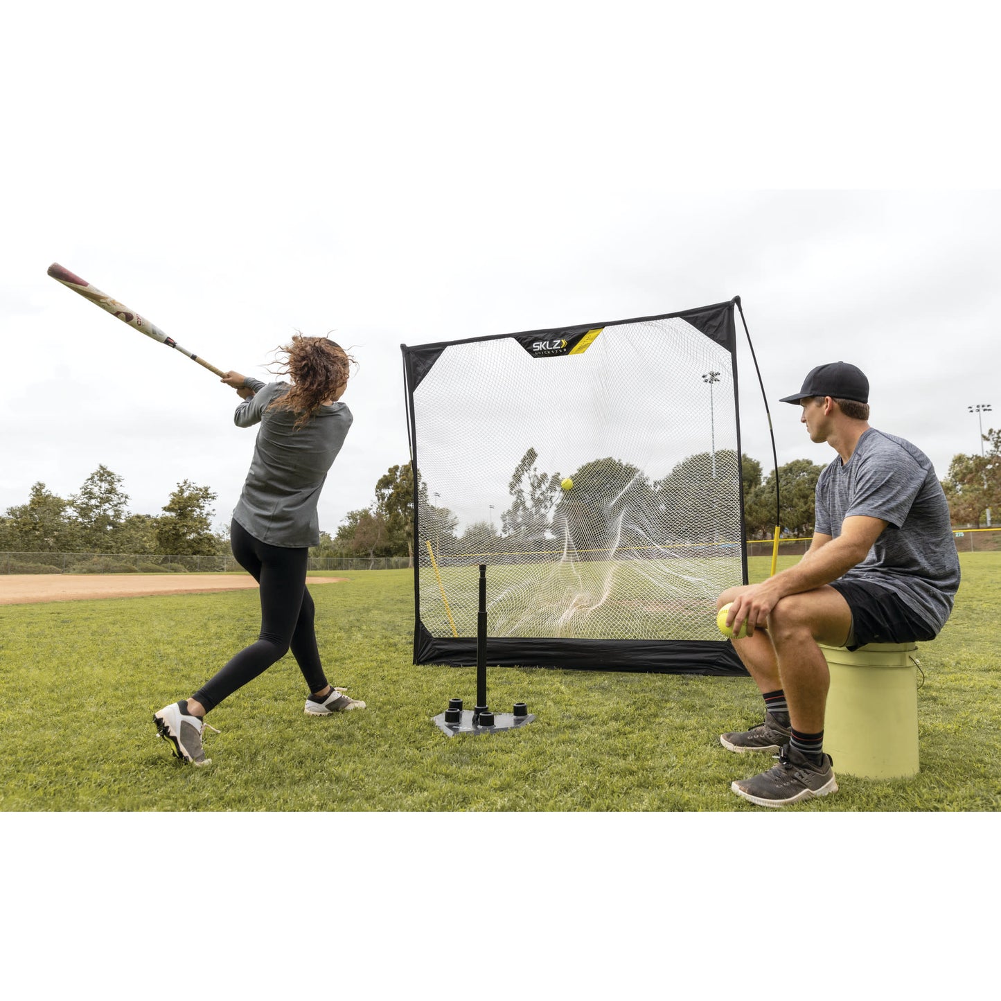 Person swings a bat at a ball in front of a portable soccer goal on a grassy field with a spectator sitting nearby