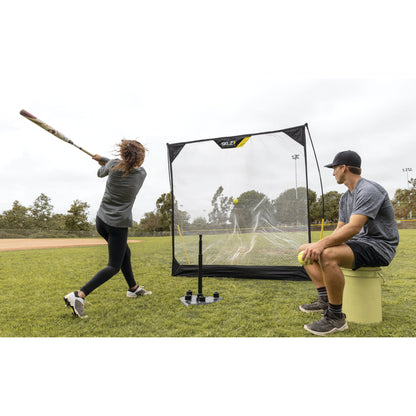 Person swings a bat at a ball in front of a portable soccer goal on a grassy field with a spectator sitting nearby