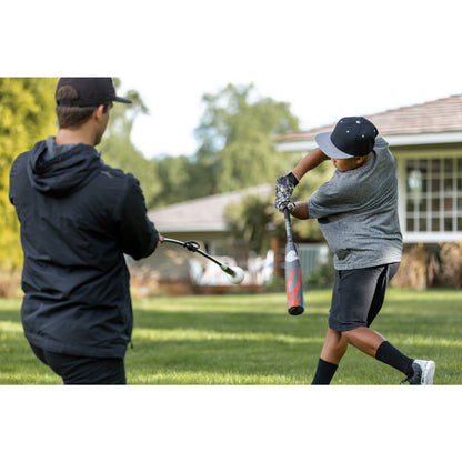 Boy swings bat while adult holds a tee outdoors on a grassy yard during daytime