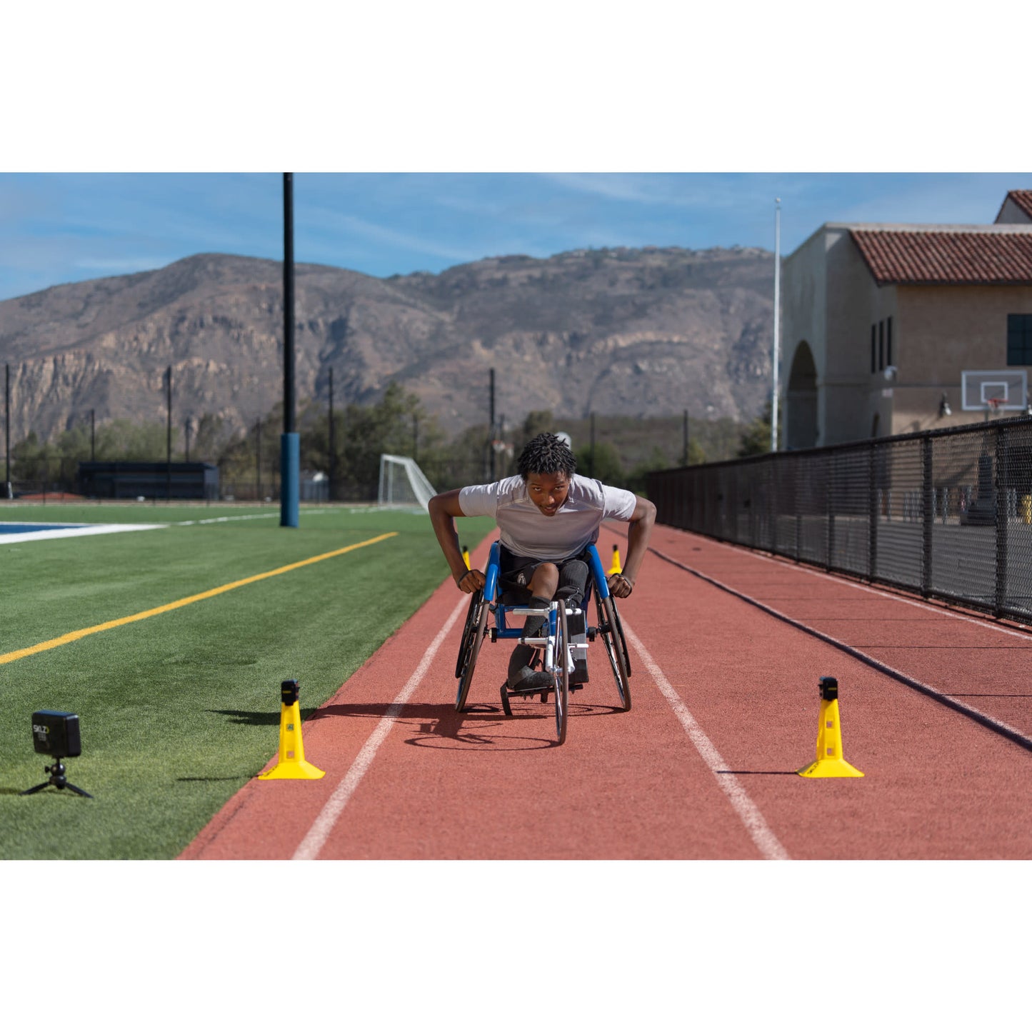 Athlete in wheelchair trains on a track with cones and cameras outdoors near a school field