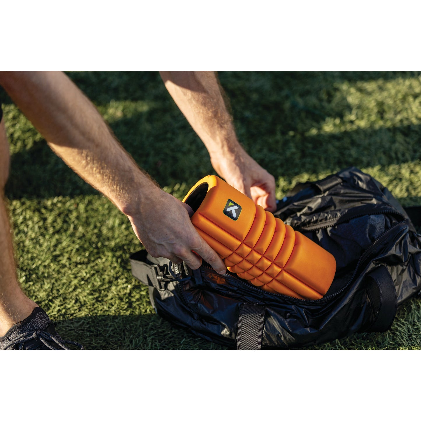 Person places orange waterproof speaker into a backpack outdoors on grassy ground.