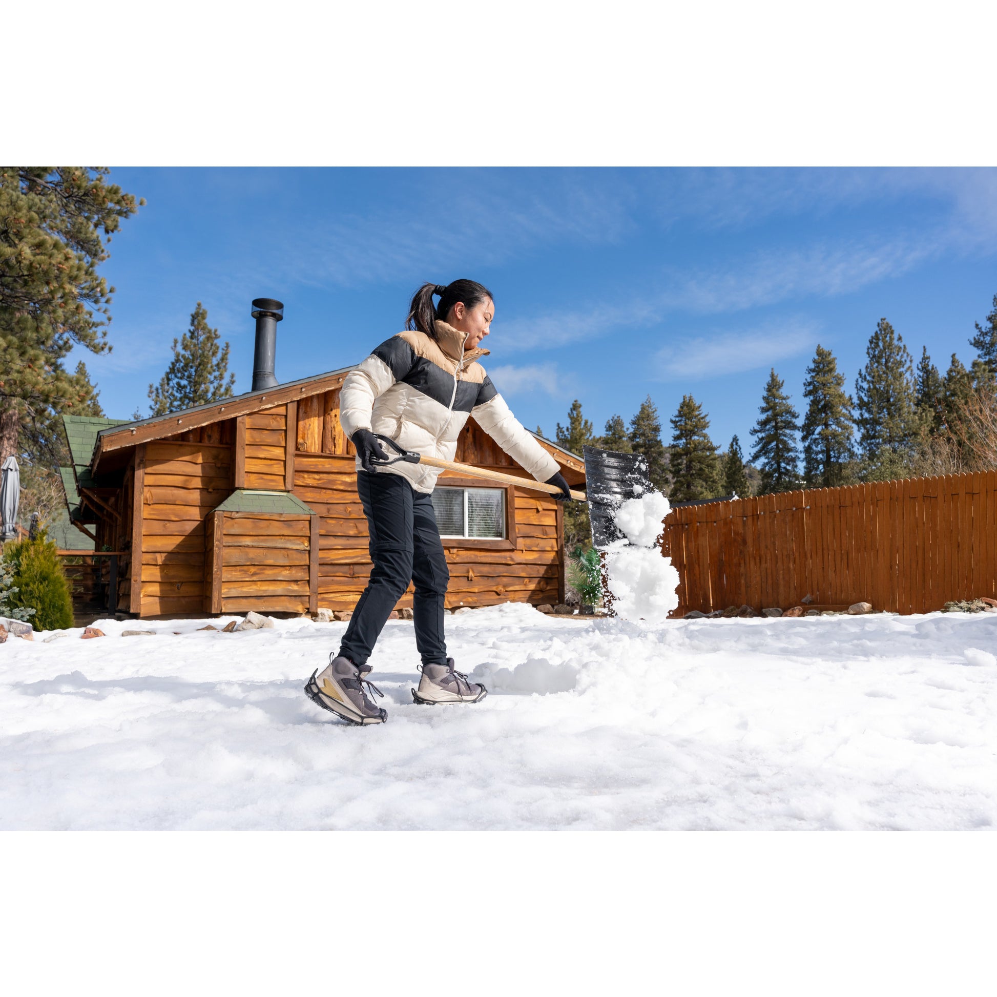 Woman shoveling snow in yard outside wooden cabin under clear blue sky
