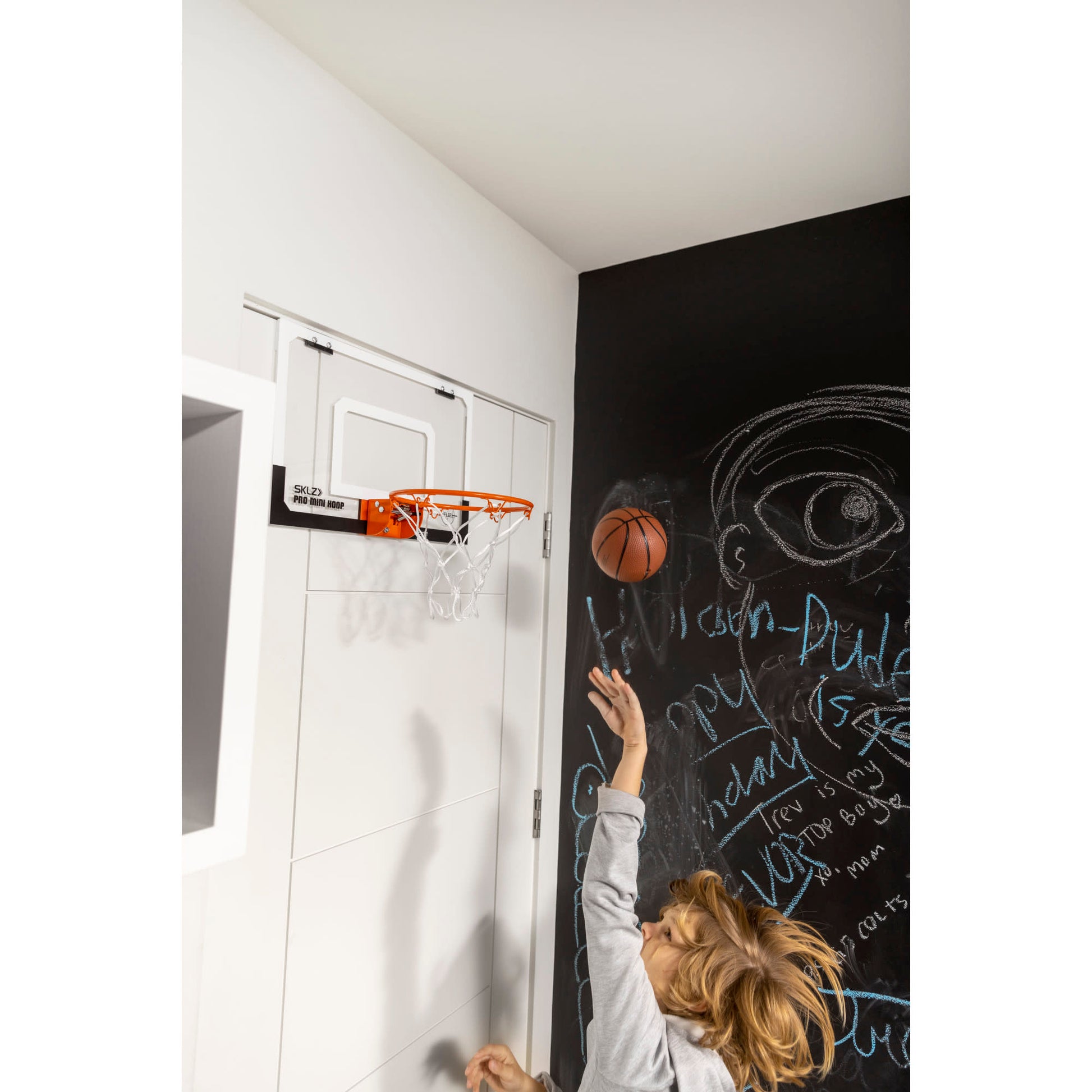 Child throws a basketball towards a hoop on a black chalkboard wall in a room.
