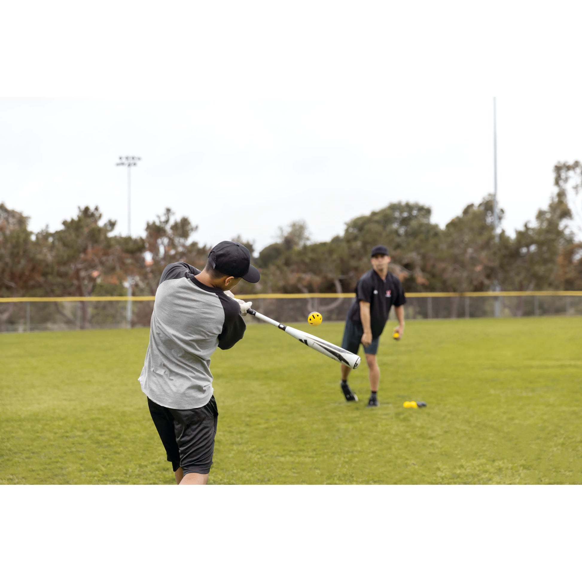 Young person practicing baseball swing on field with coach nearby in the background