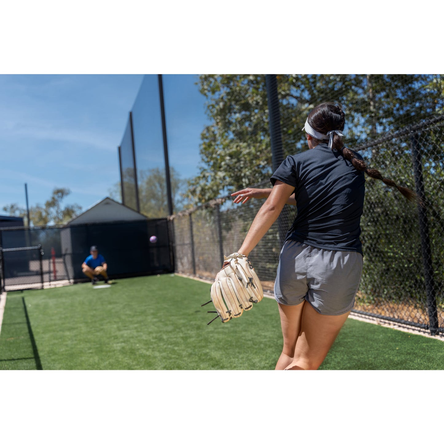 Girl throws a ball at a net in a fenced outdoor sports area on a sunny day.