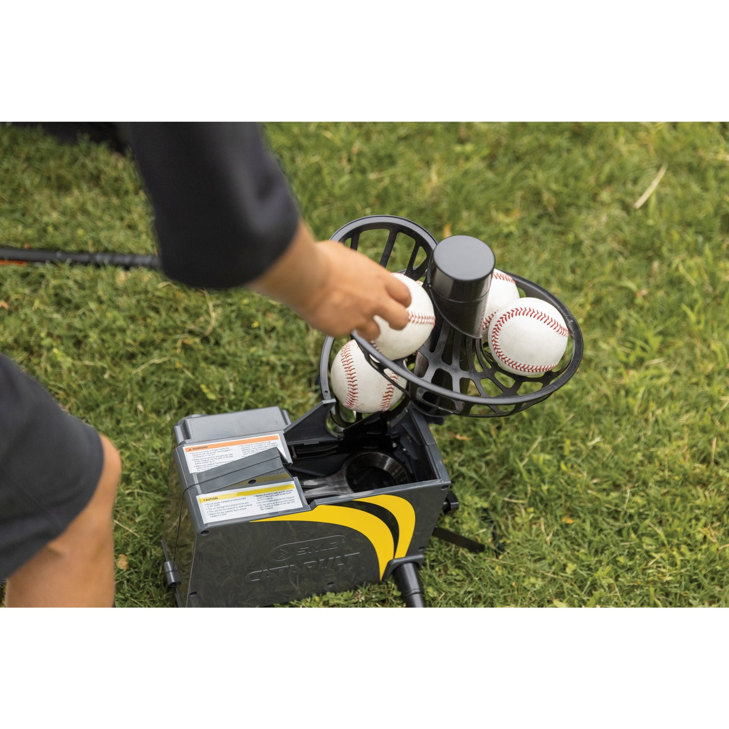 Person loads baseballs into a pitching machine on green grass field.