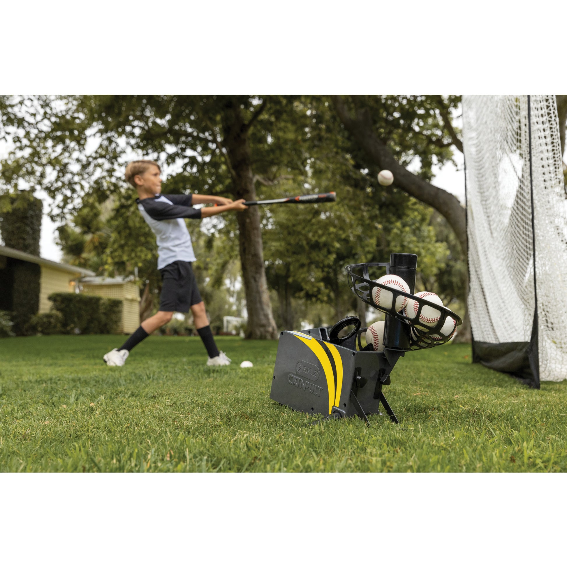 Girl practices baseball batting with a bat near a ball net on a grassy field.