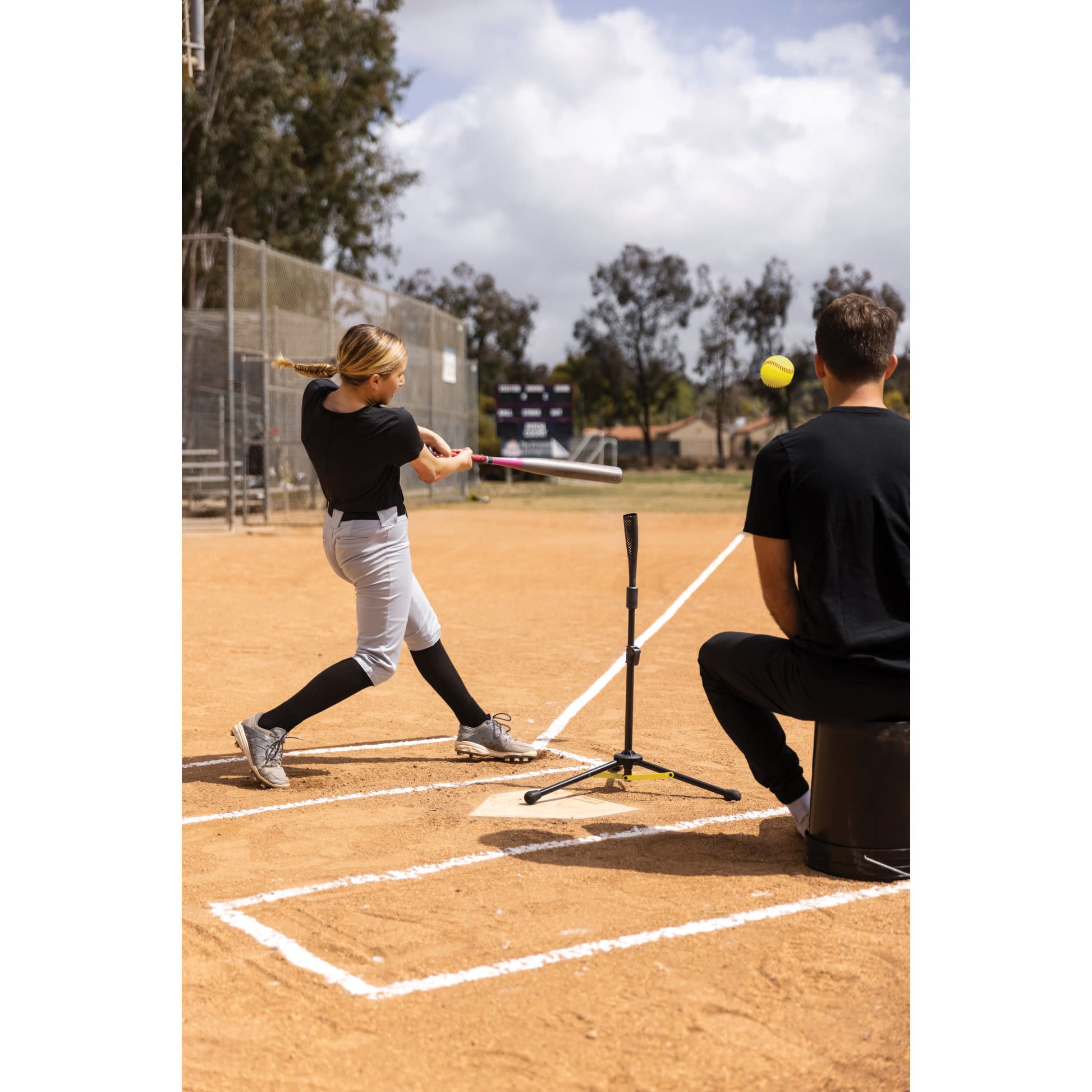Girl swings bat to hit ball during practice on baseball field with coach nearby