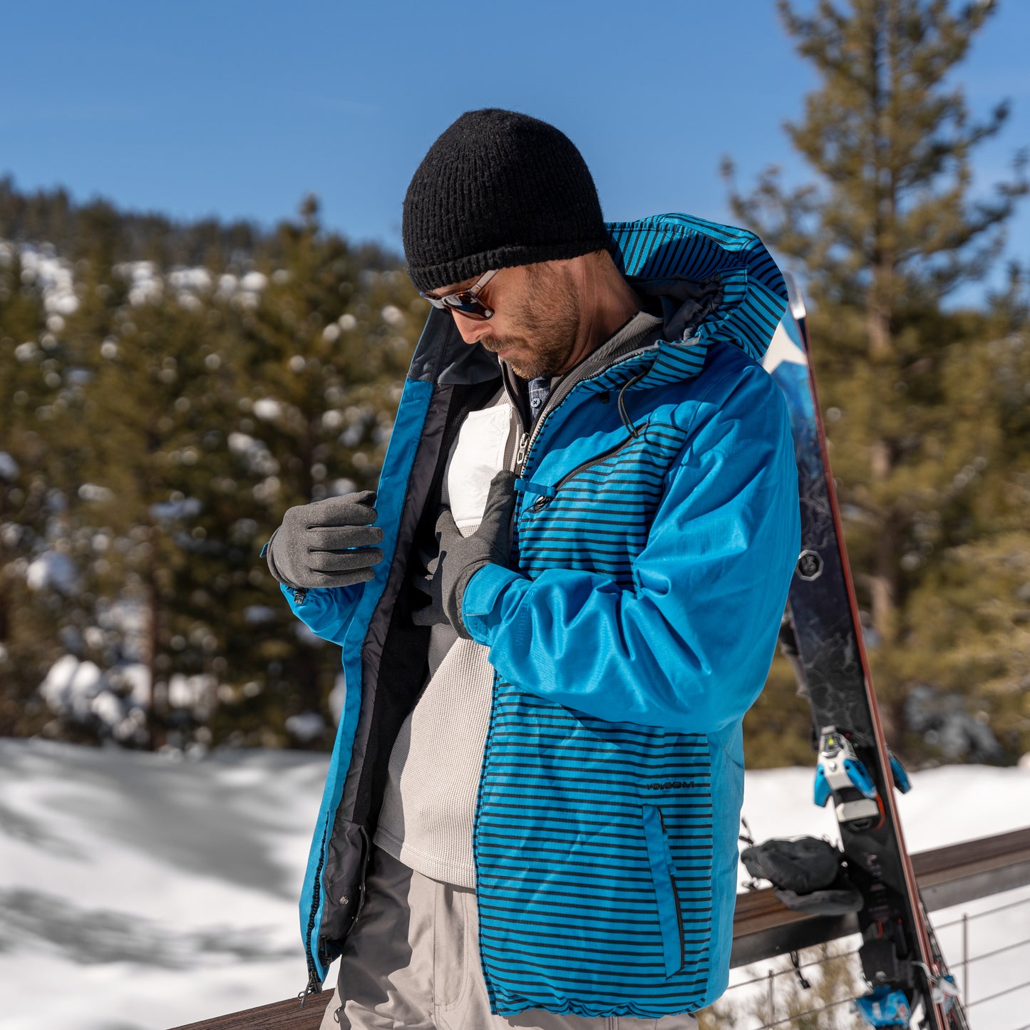Man adjusts jacket in snowy outdoor environment with skis on shoulder