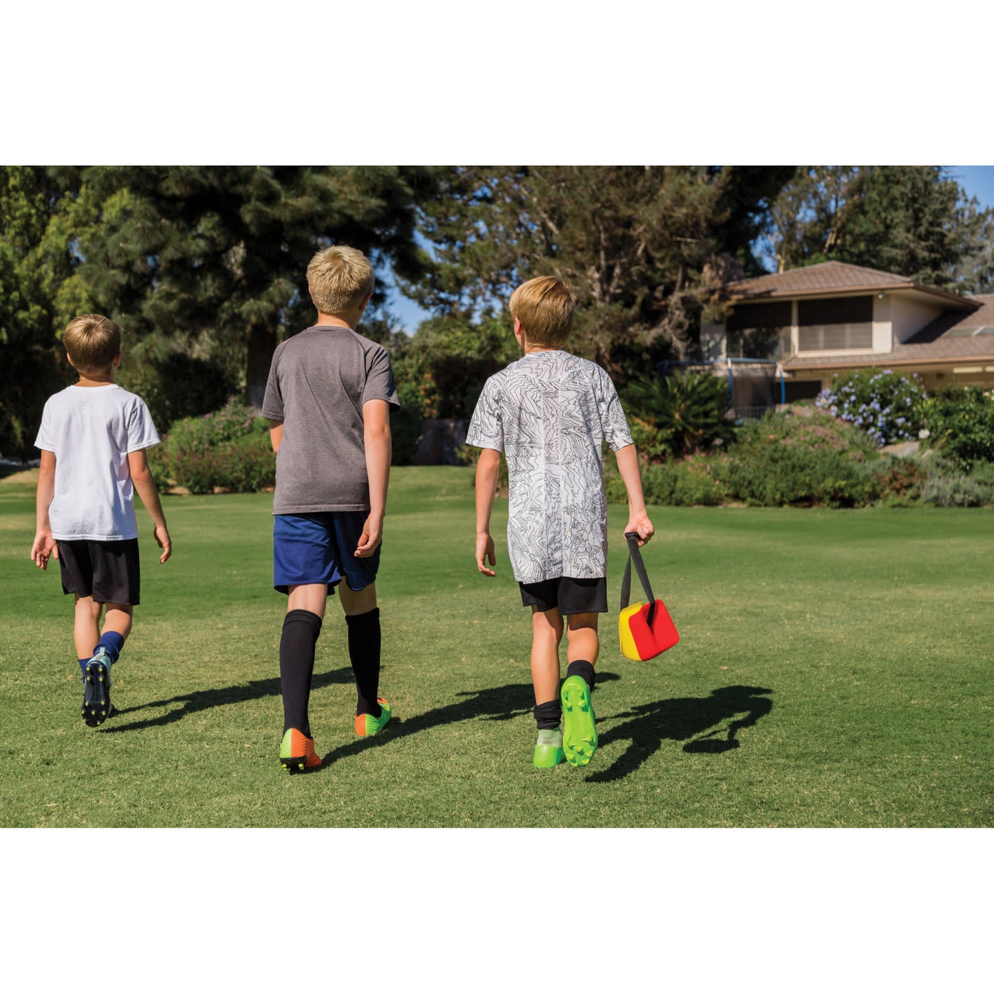 Three children walk on a grassy field carrying sports equipment in a park with trees and a building
