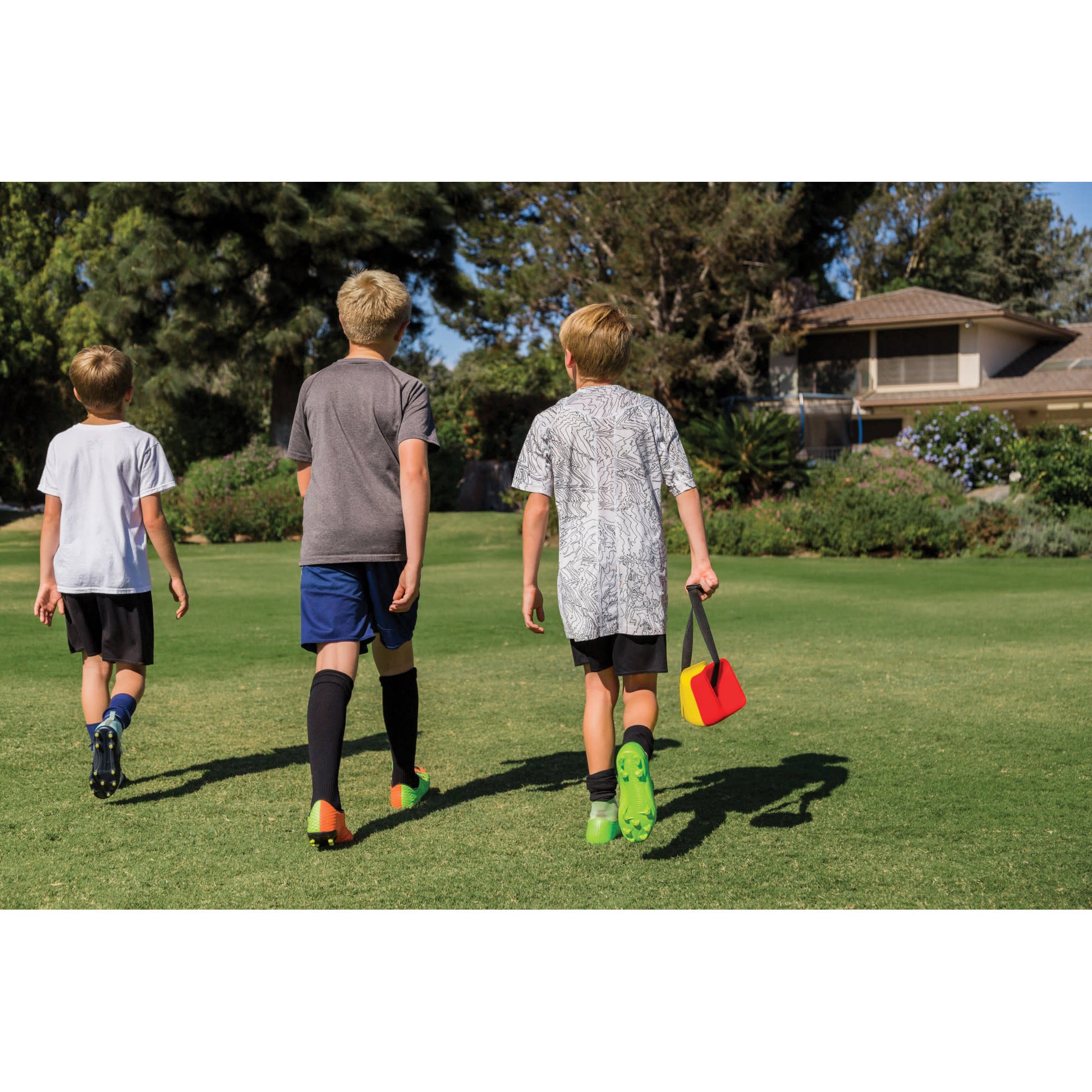 Three children walk on a grassy field carrying sports equipment in a park with trees and a building