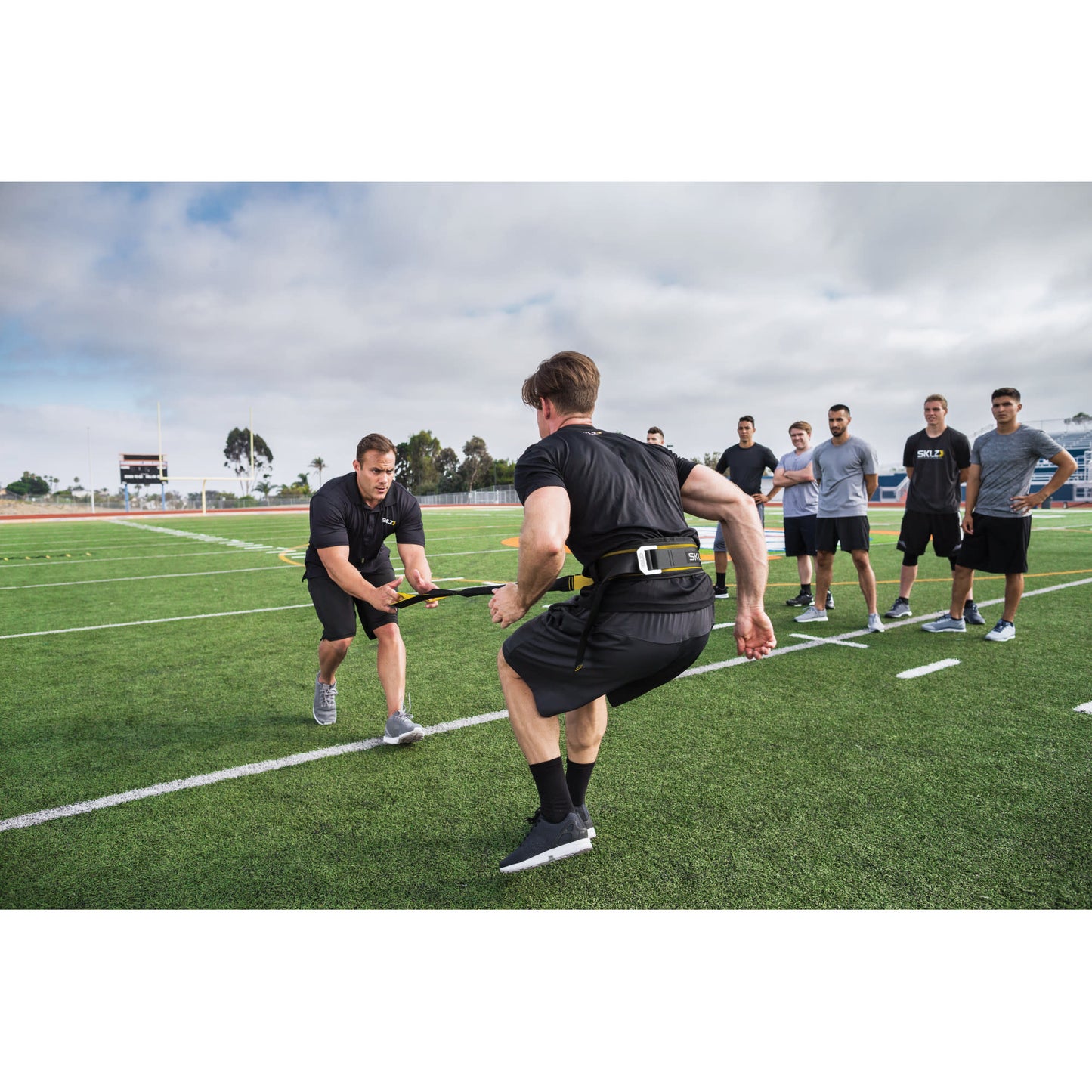 Male athlete using SKLZ Acceleration Trainer with a Coach on a football field with team on sideline
