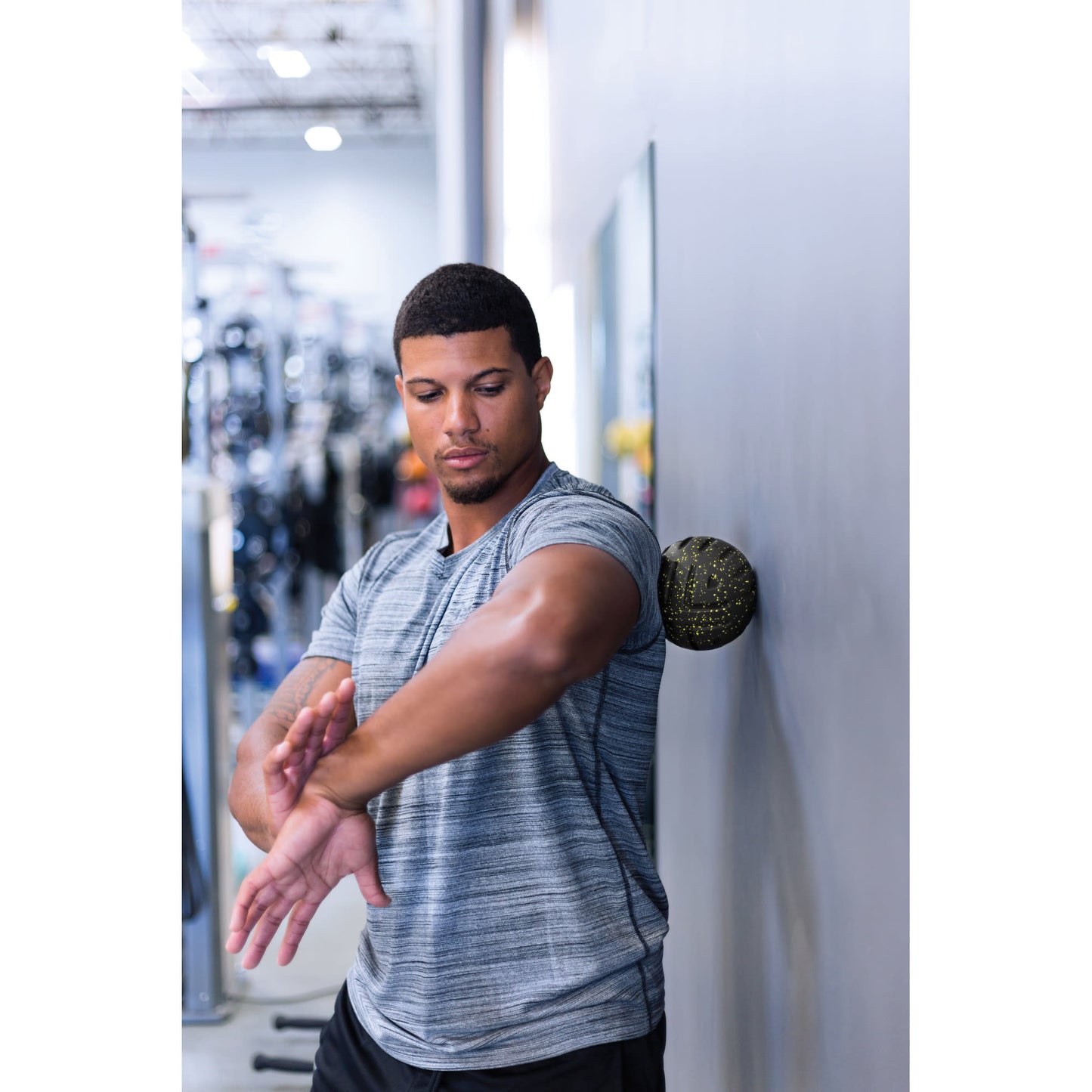 Man stretches arm against wall with a therapy ball behind him in a gym environment