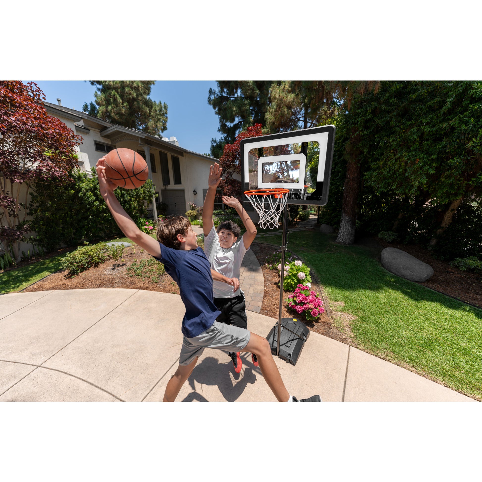 Two children play basketball on an outdoor court near a house and garden
