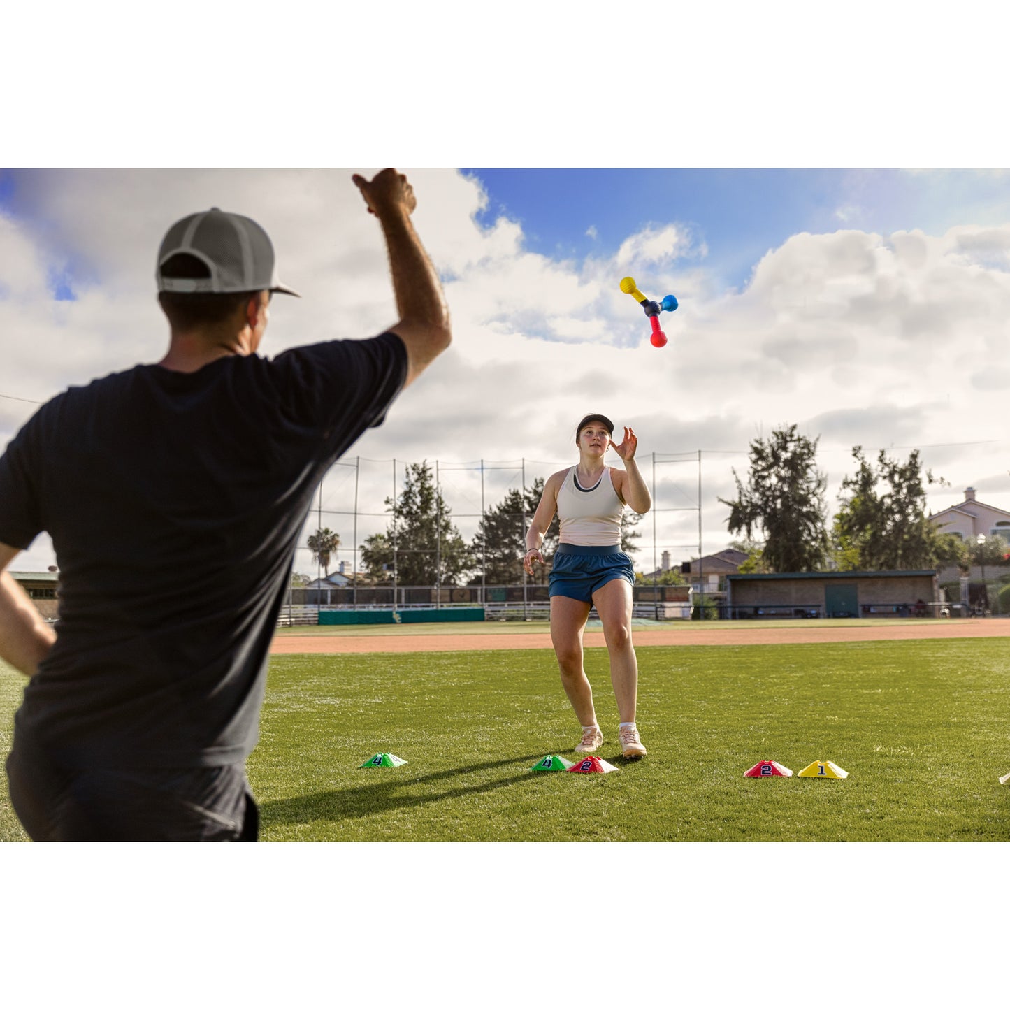 Woman throws pins at boy during outdoor game on baseball field with cones and cloudy sky