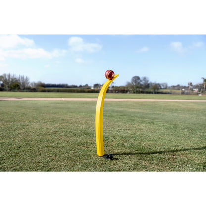 A curved yellow cricket training aid holds a ball on a grassy field under a clear blue sky.