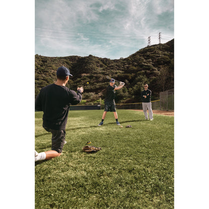 Three people practice baseball on a grassy field with a hilly landscape and power lines in the background