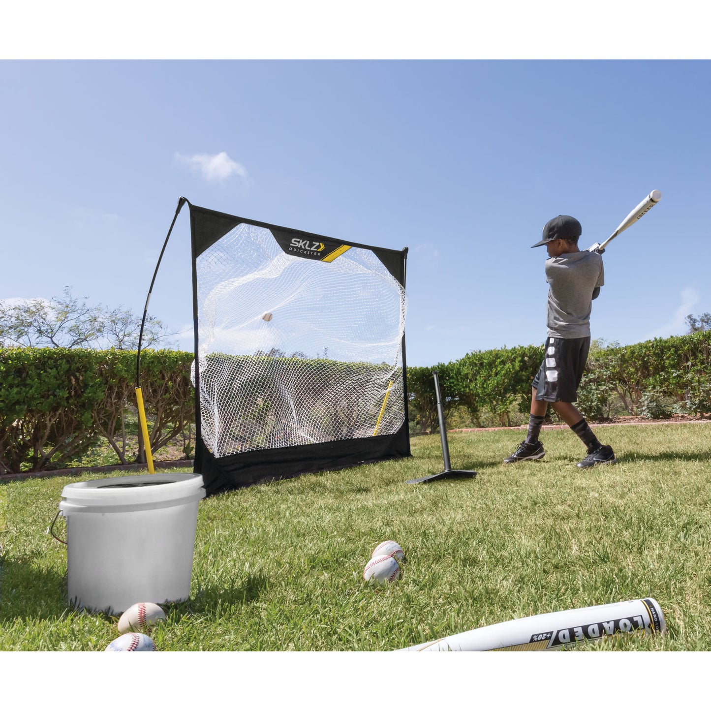 Child practices baseball hitting with a bat near a net on a grassy field under a blue sky