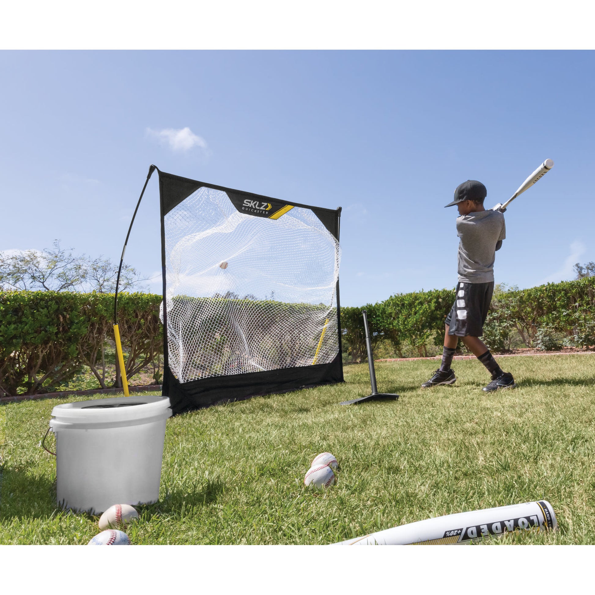 Child practices baseball hitting with a bat near a net on a grassy field under a blue sky