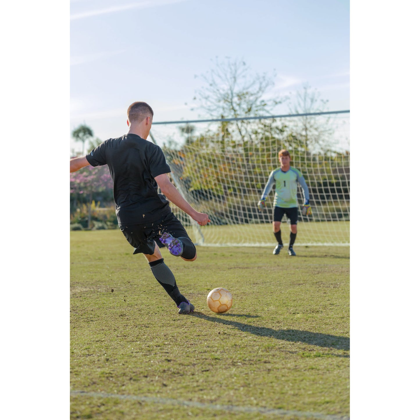 Player kicks soccer ball towards goalkeeper on a grassy field during daytime