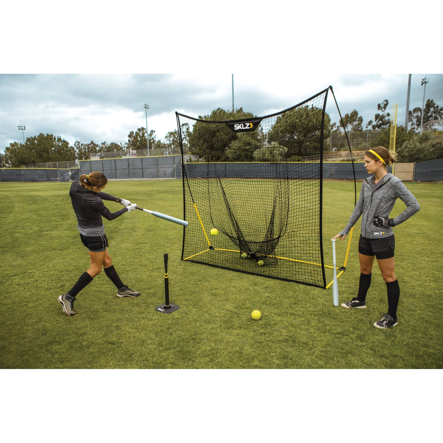 Two women practice softball hitting with a tee on a grassy field near a protective net