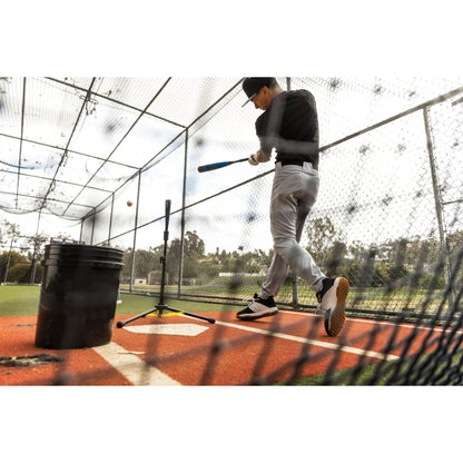 Baseball player swings at ball during practice inside a netted batting cage on a field.