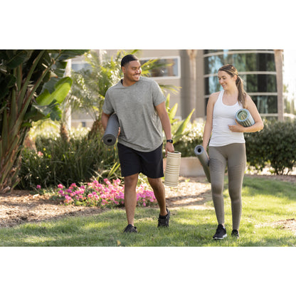 A man and woman walk outdoors carrying yoga mats and a water bottle in a sunny garden area.