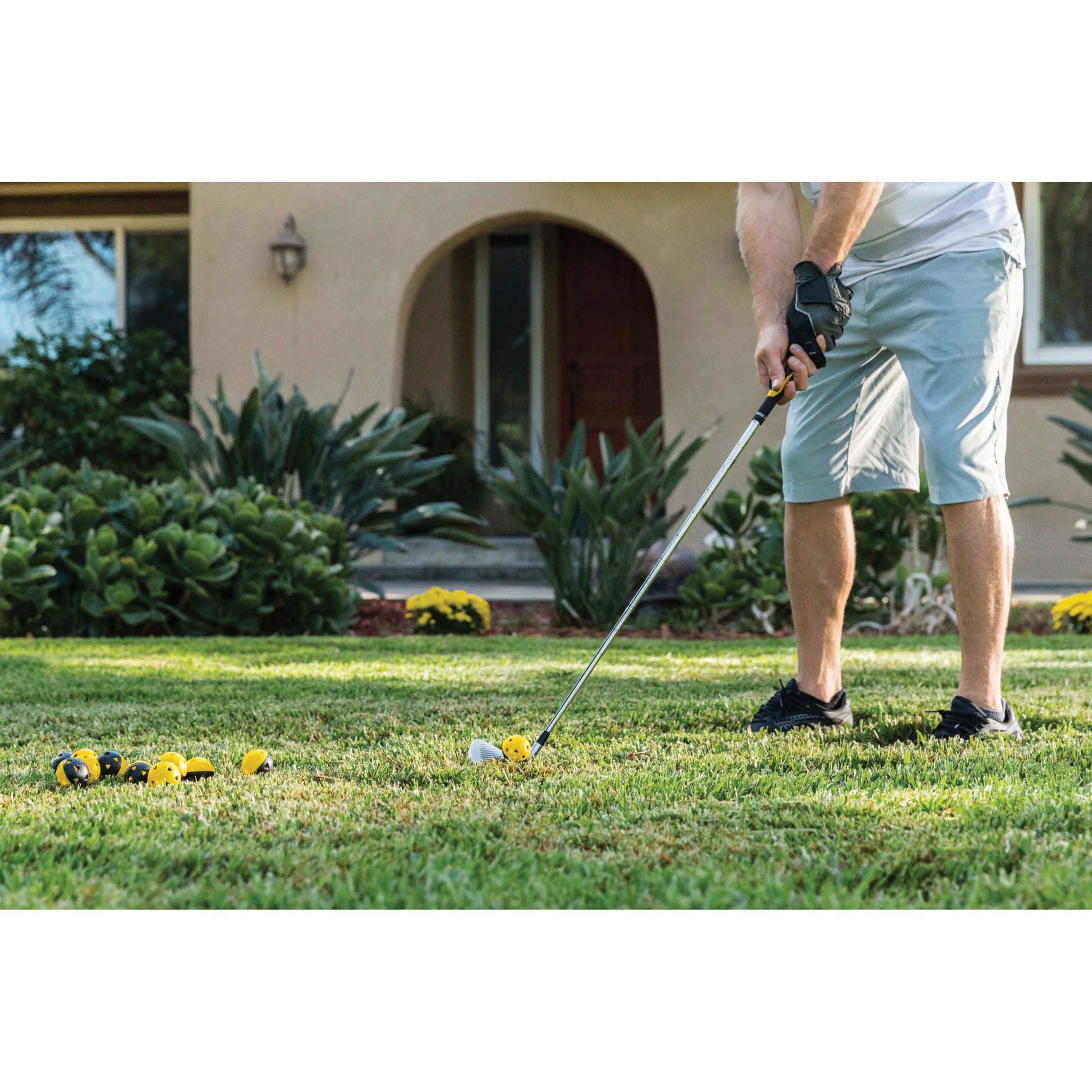 Person practicing golf on a lawn with golf balls and clubs near a house yard