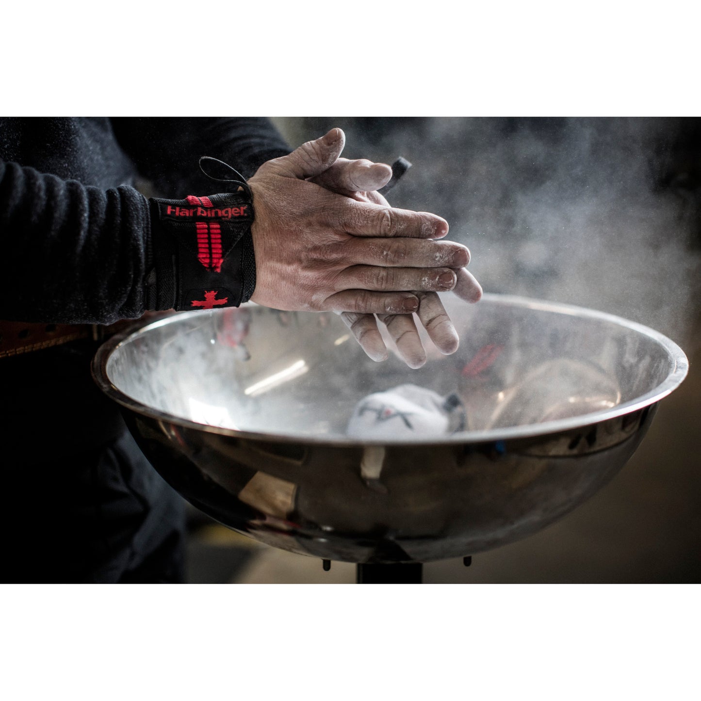 Hands clap above a metal bowl, releasing white chalk dust in a gym setting.