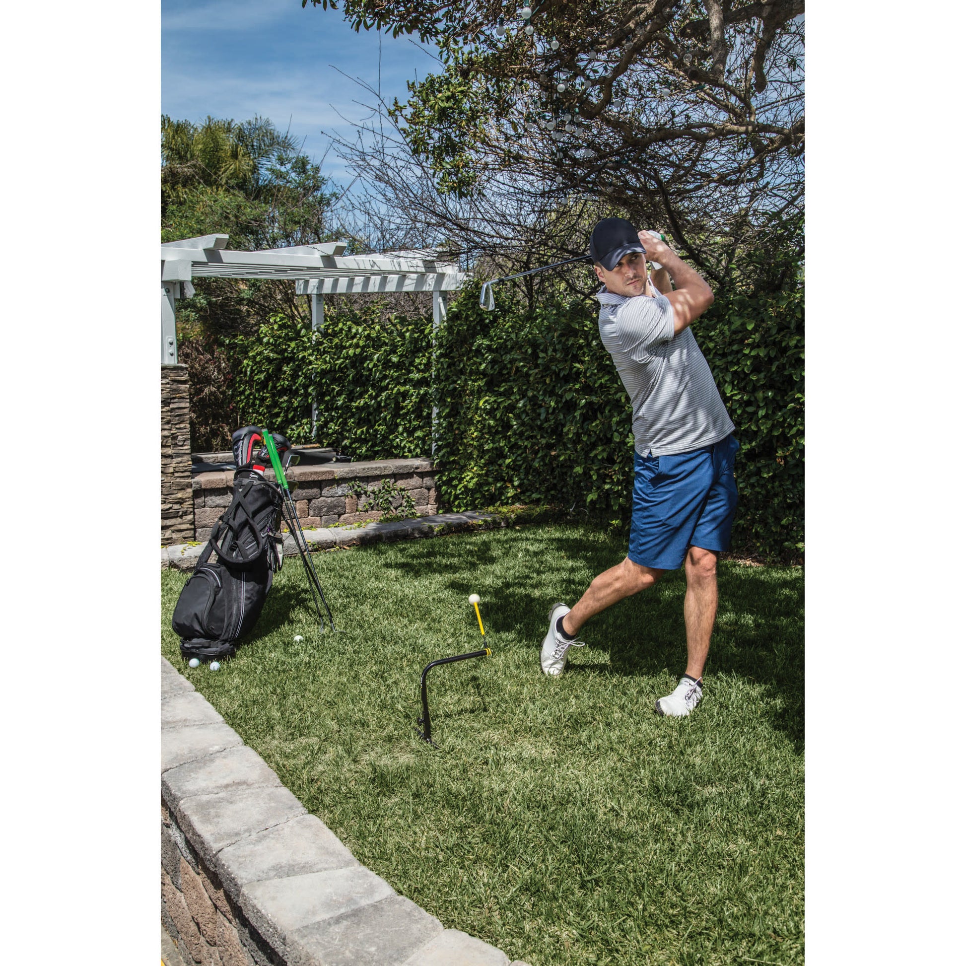Golfer swings club in backyard with grass tree and golf bag nearby