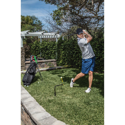 Golfer swings club in backyard with grass tree and golf bag nearby