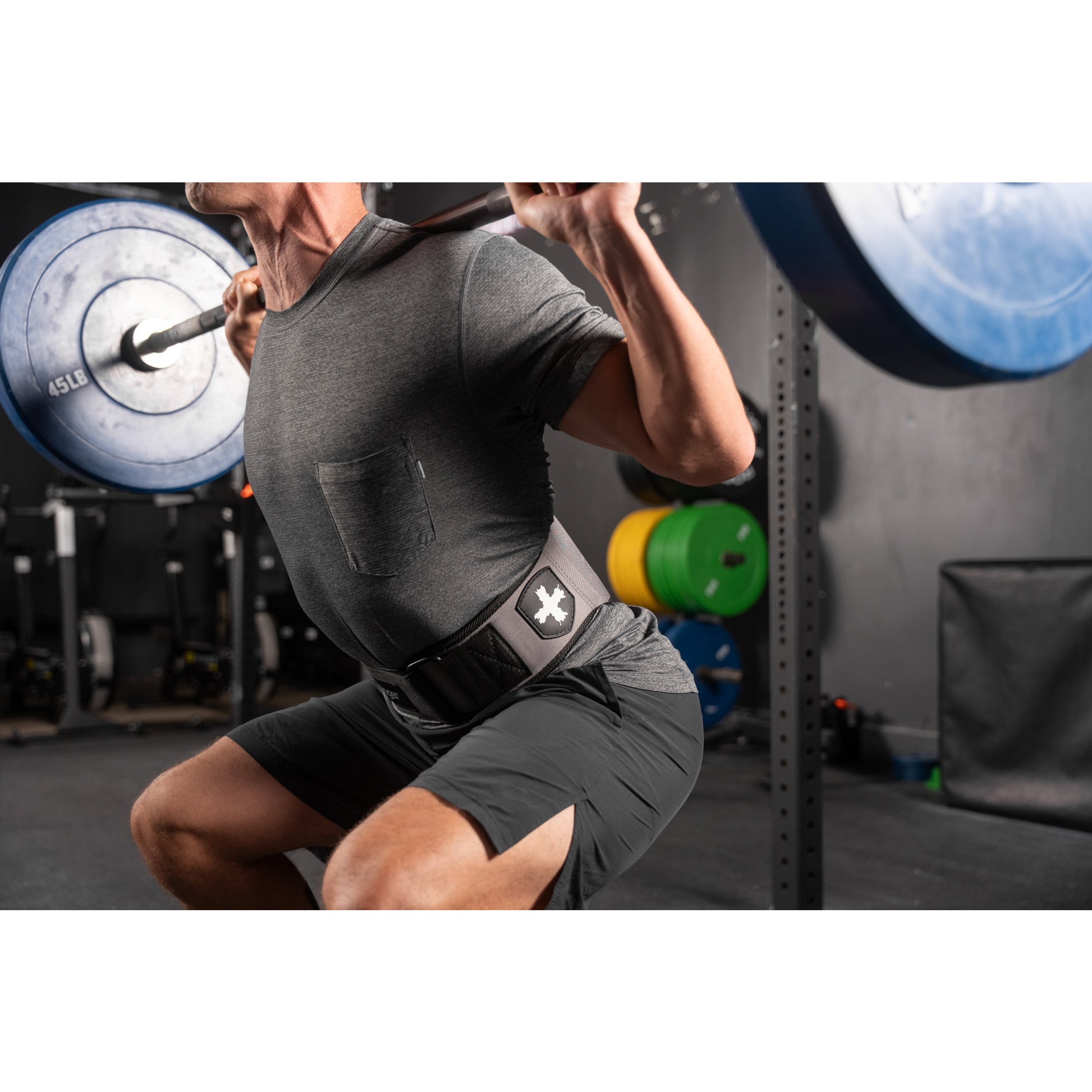 Man squatting with barbell in gym, wearing a lifting belt near weight racks.
