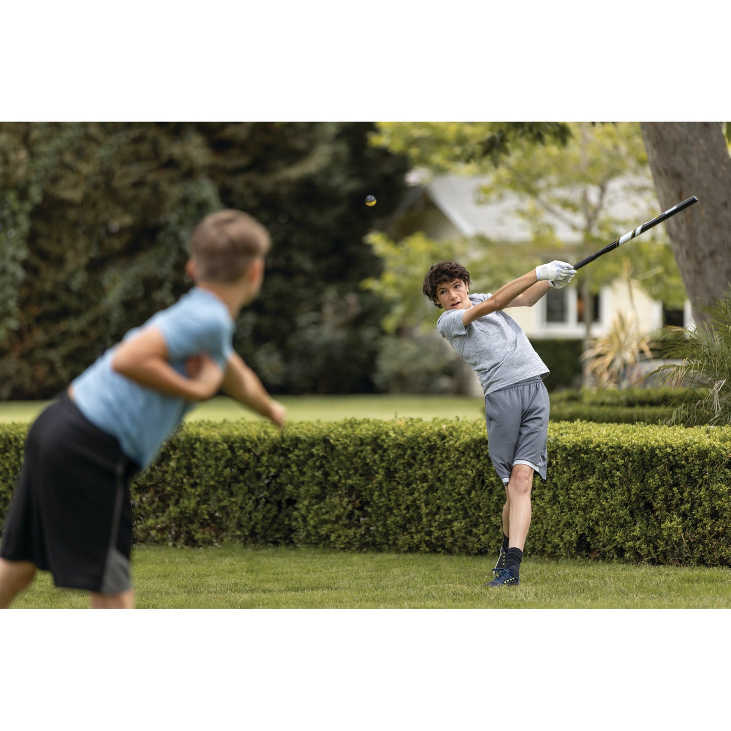 Boy swings golf club in garden while another boy watches nearby