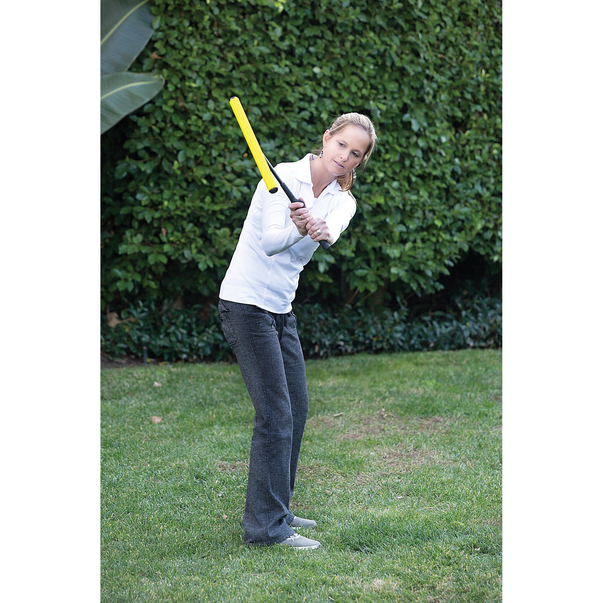 Woman swings a yellow golf club on a grassy lawn with a bushy background
