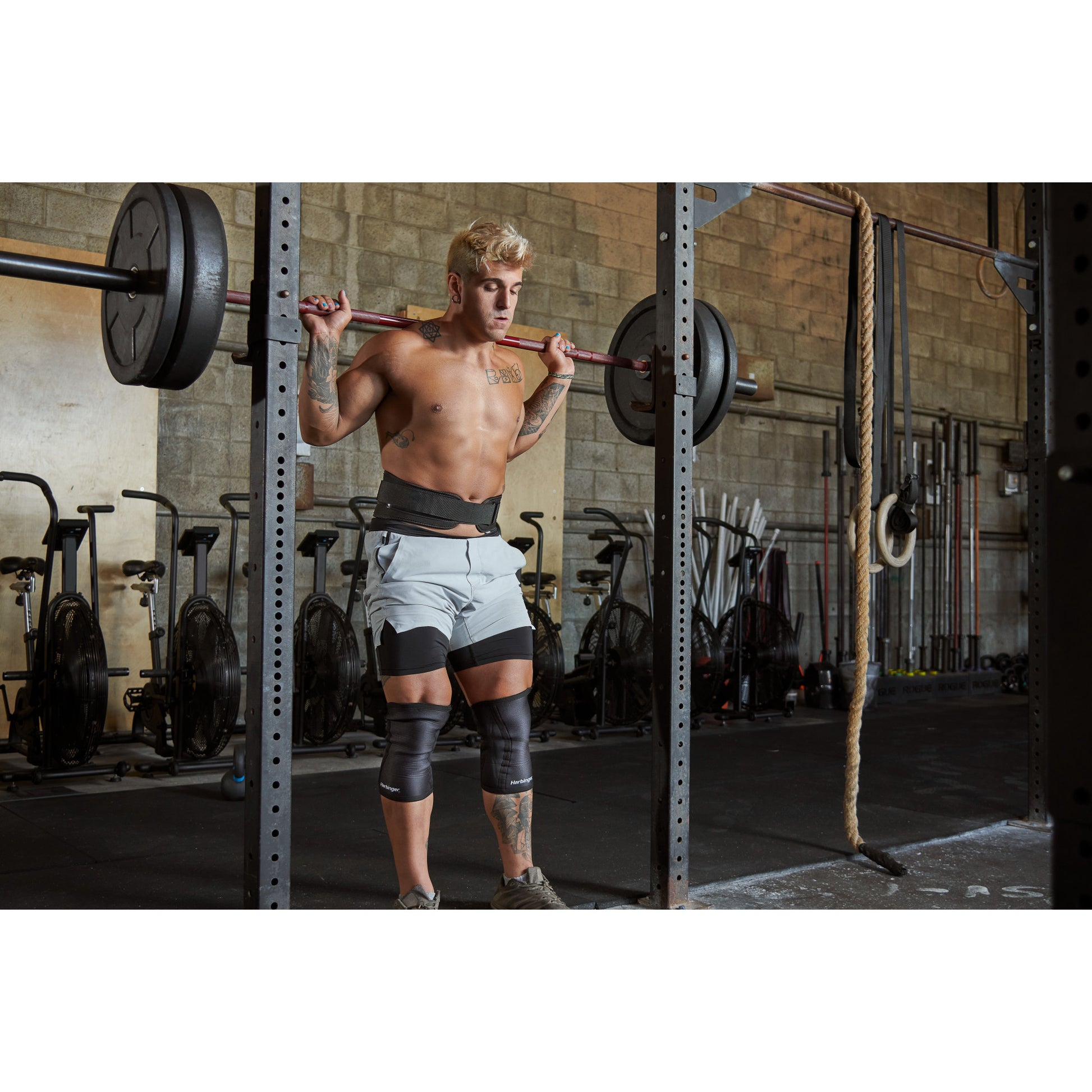 Man lifting barbell weights in gym, wearing knee supports, surrounded by exercise equipment.
