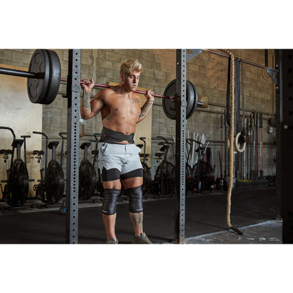 Man lifting barbell weights in gym, wearing knee supports, surrounded by exercise equipment.