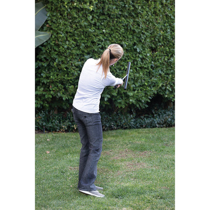 Woman practices golf swing outdoors on grass with green foliage in the background