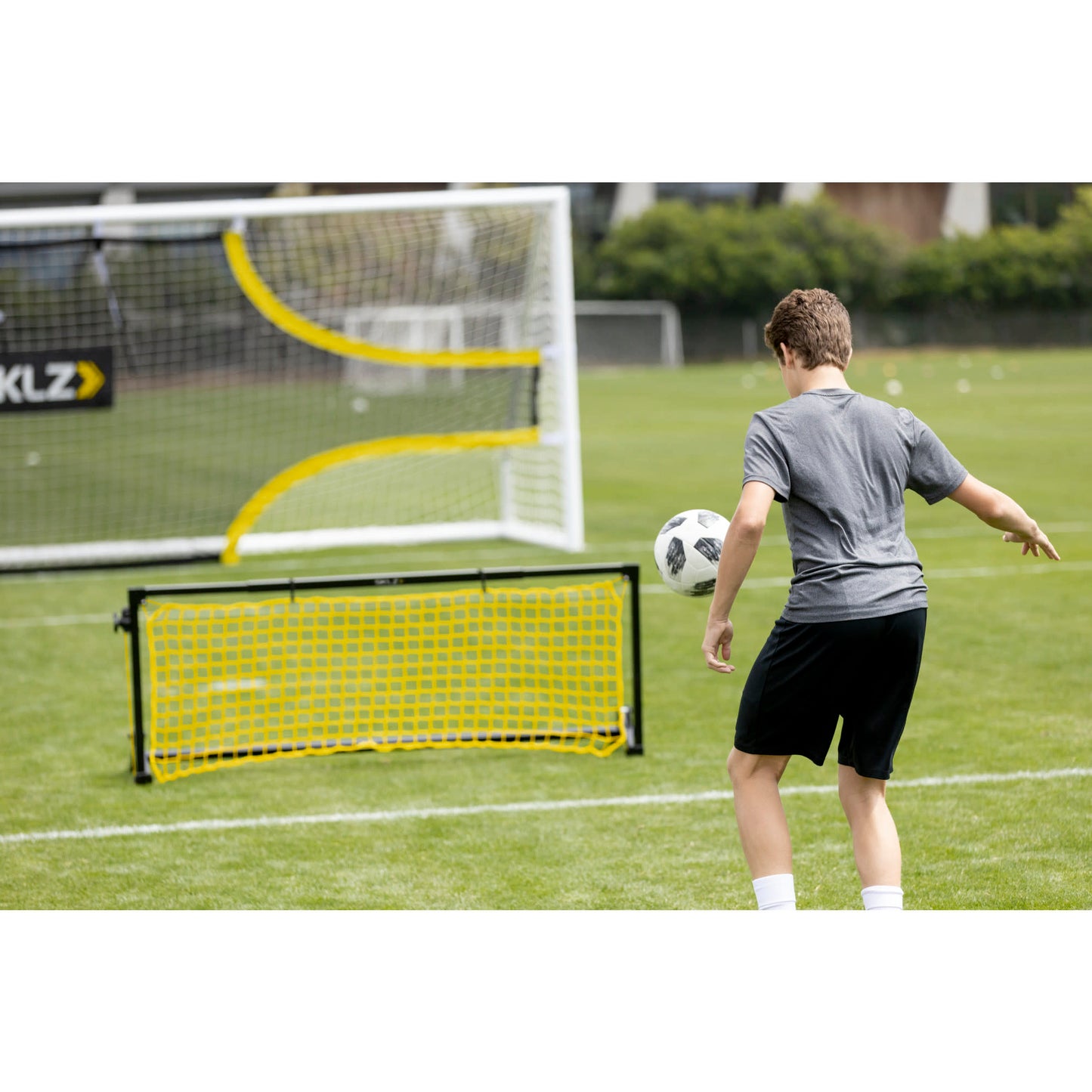 Boy practices soccer shooting on goal net with soccer ball on a grassy field.