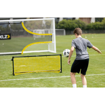 Boy practices soccer shooting on goal net with soccer ball on a grassy field.