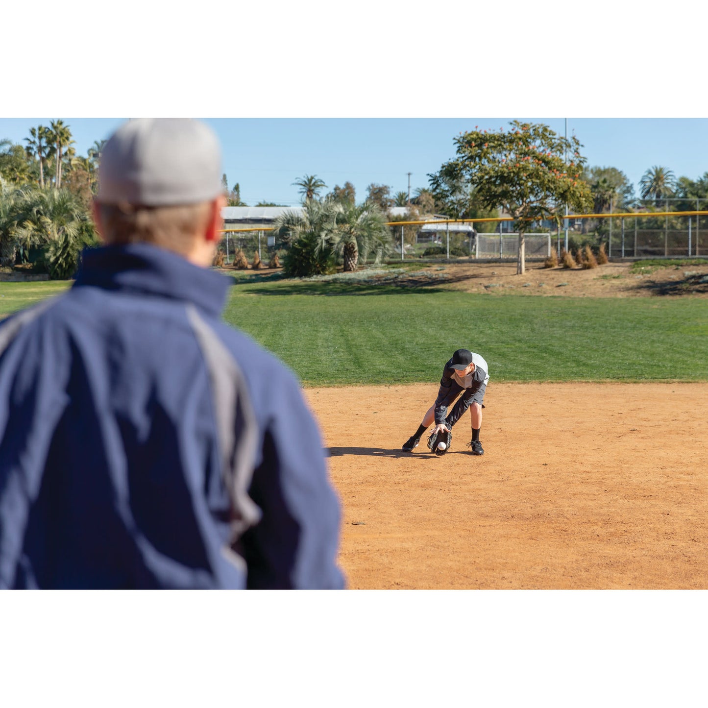Child fields a baseball on a dirt infield during a game with a coach observing nearby
