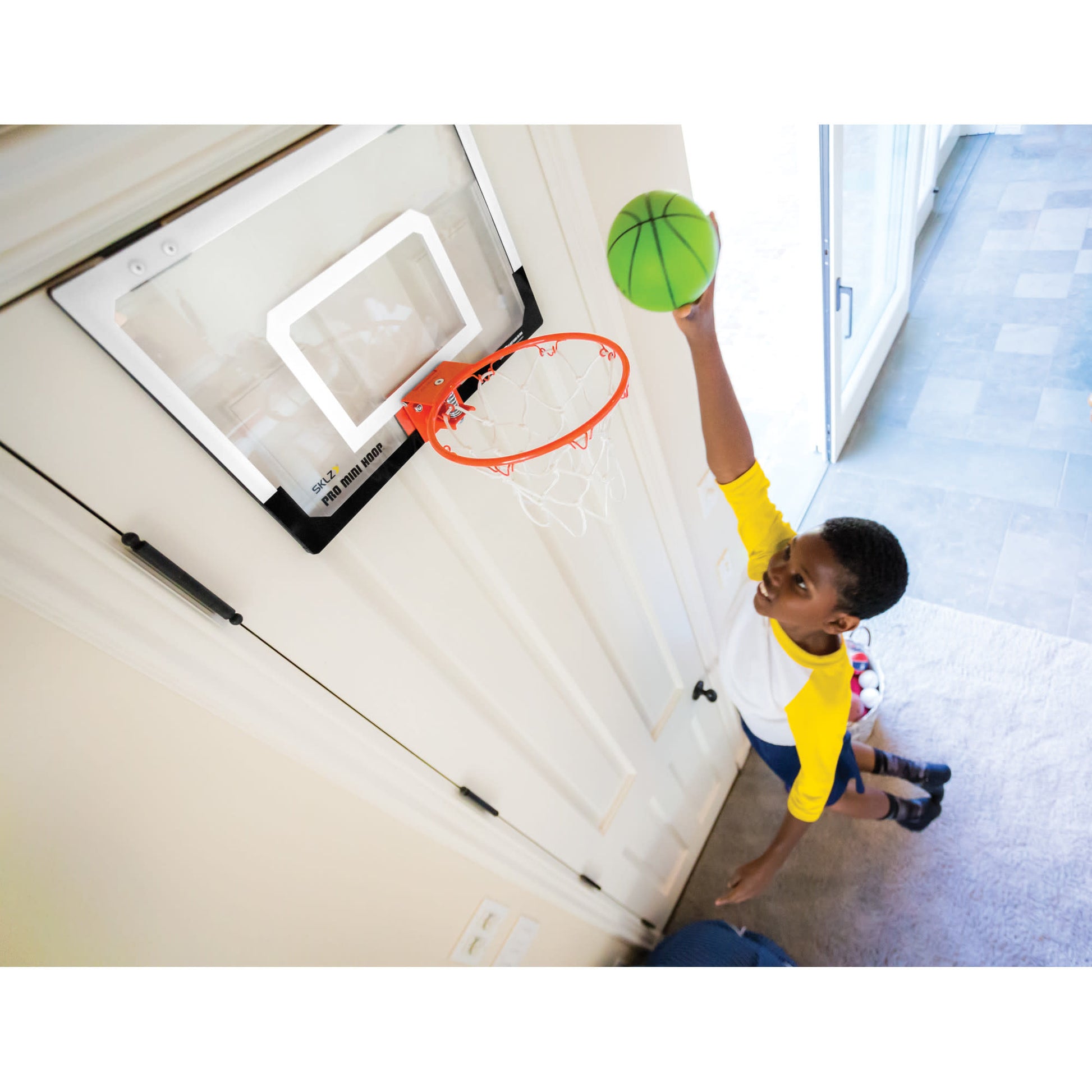 Boy reaches for basketball near indoor hoop in a room with light-colored walls and flooring.