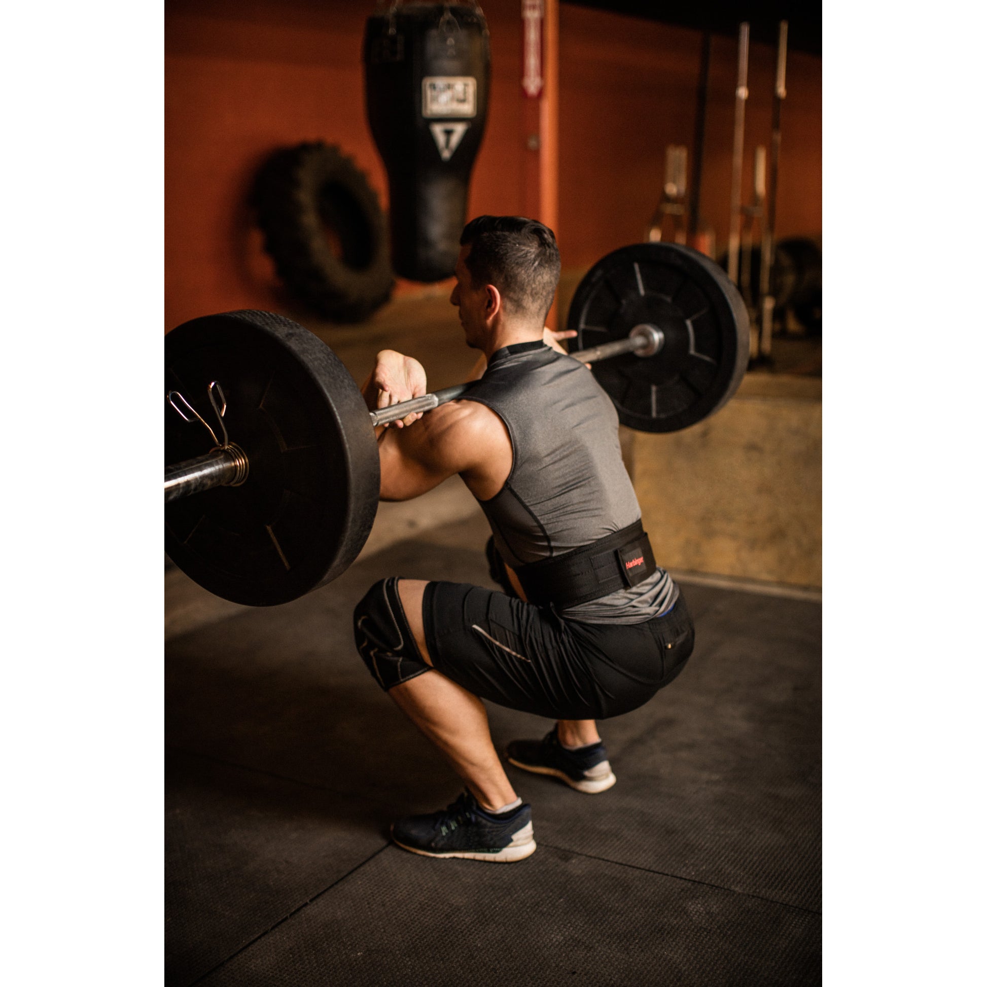 Man squats with barbell in gym, surrounded by weights and equipment.