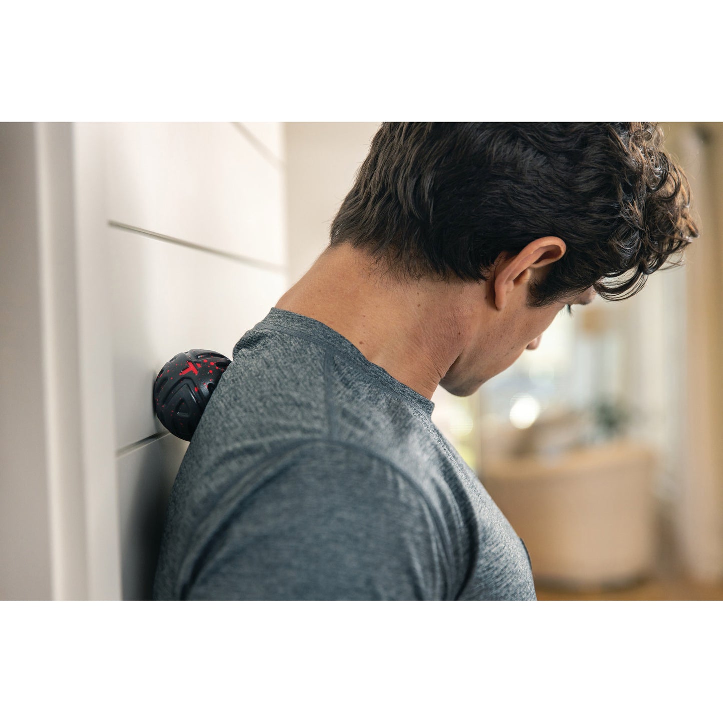 A man leans against a wall with a massage ball placed between his back and the wall in a room.