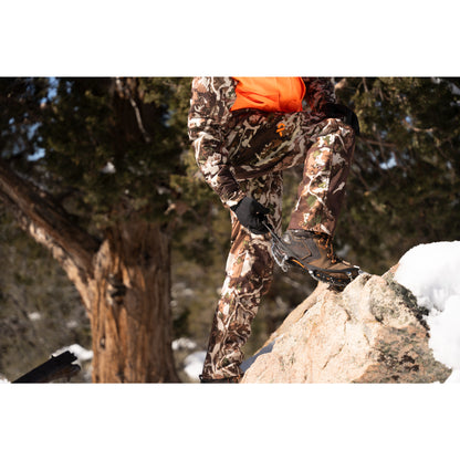 Person in camouflage clothing and orange jacket plants a snowshoe on a rock outdoor in winter