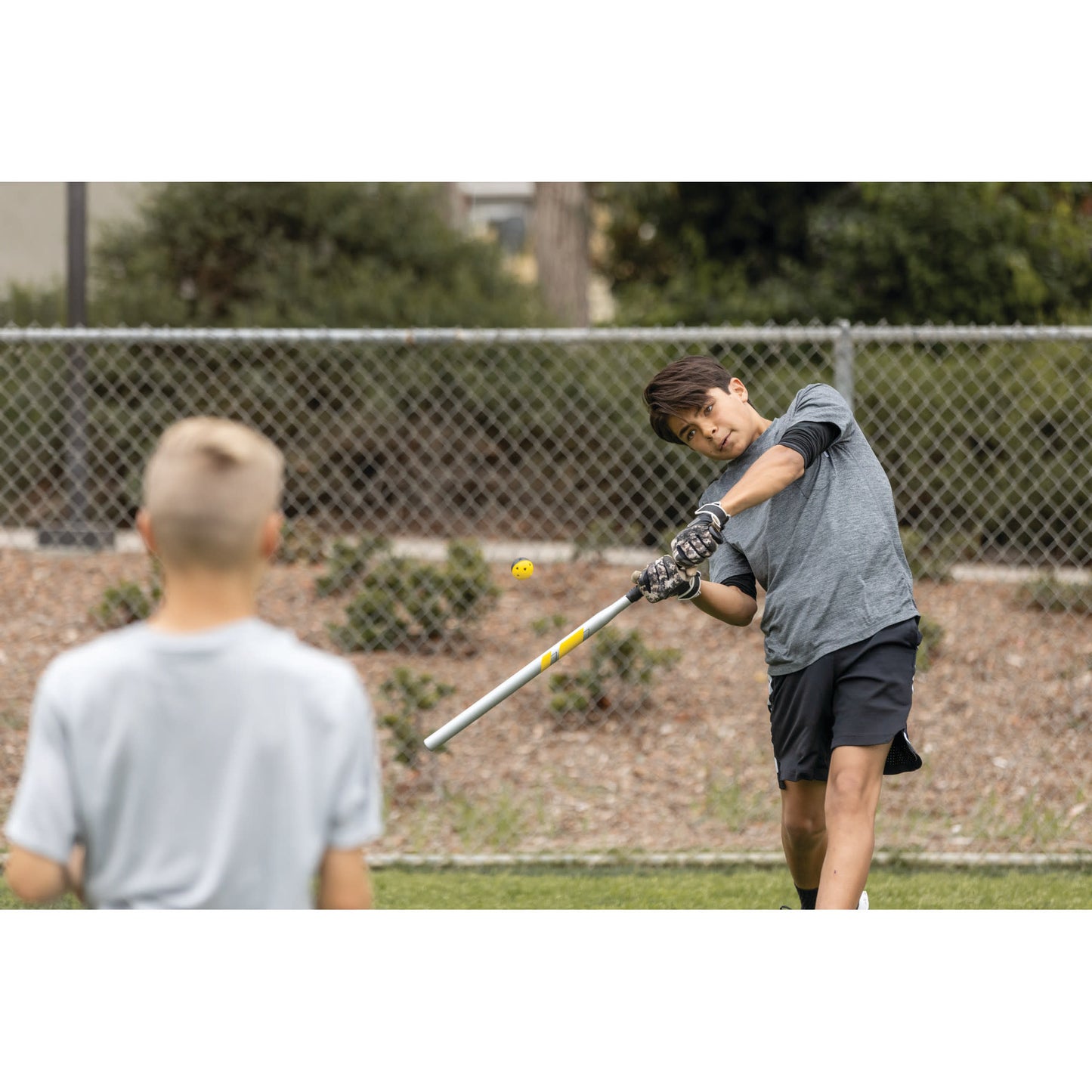 Boy hits a golf ball with a club on a grassy field near a fence in a park