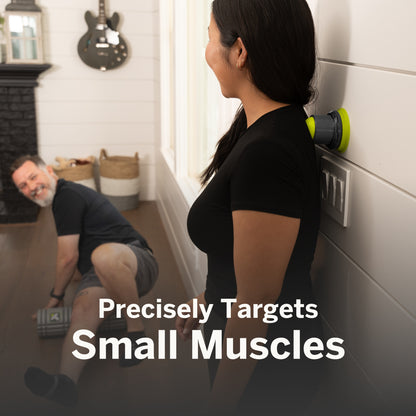 Woman uses a muscle stimulator on her back while man exercises on the floor indoors.